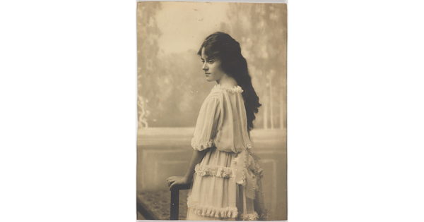 A black-and-white photograph of a young White woman with long dark hair wearing a ruffled dress standing back to the camera but head turned in profile.