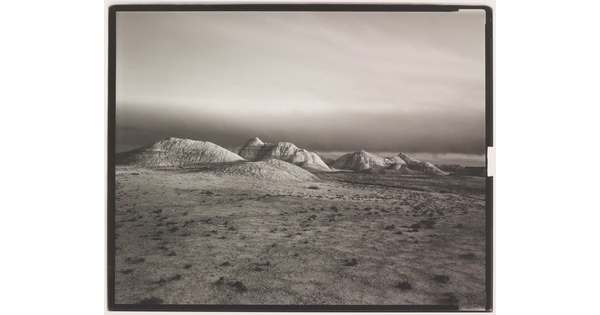 A black-and-white landscape photograph of distant hills at the edge of a flat desert under a dark sky.