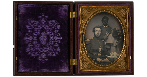 A black-and-white photograph in a hinged case with an oval frame of a seated White man in a military uniform and a  young Black boy standing behind him.