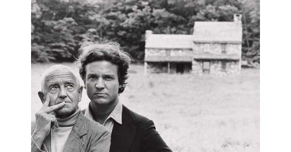 A black-and-white photograph of two White men, one older and one younger, standing in front of a stone farmhouse in the distance.