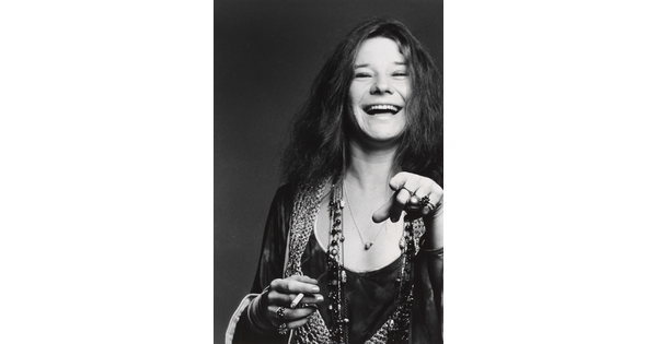 A black-and-white portrait photograph of a White woman with long hair, an open-mouthed smile, holding a cigarette in one hand and pointing at the camera with the other.