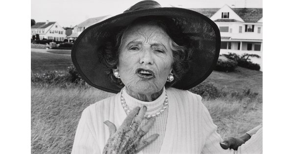 A black-and-white photograph of an elderly White woman wearing a wide-brimmed sunhat, pearl earrings and necklace, looking as if she is speaking.