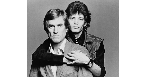 A black-and-white studio portrait photograph of two White men, one standing behind the other with his arms wrapped around the other&#039;s shoulders.