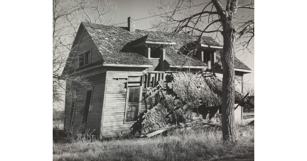 A black-and-white photograph of an abandoned, two-story, wood home that is falling apart.