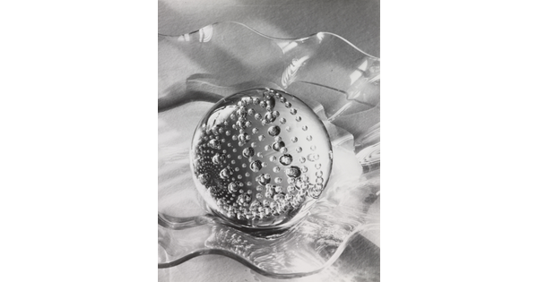 A black-and-white still life photograph of a clear glass, circular paperweight filled with bubbles in a clear, wavy glass bowl on a table.