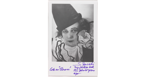 A black-and-white photograph of a woman wearing blotchy white face paint, a tall black hat, and frilly white collar, above a handwritten note.