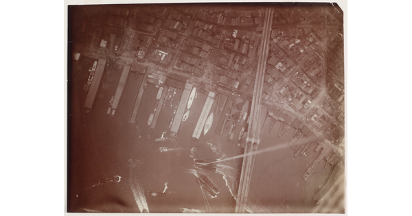 A black-and-white photograph of an aerial view of a boat harbor next to a city and a long bridge crossing the water into the city.