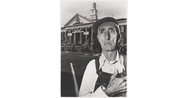 A black-and-white photograph of an elderly White man wearing a baseball cap, overalls, hand over his heart standing in front of a courthouse decorated in flags and buntings.