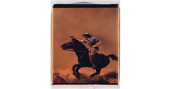 A color photograph of a White cowboy on a galloping horse, in profile, pointing a revolver in front of him.