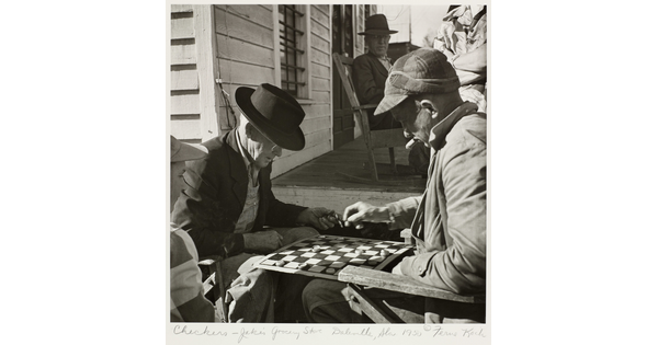 A black-and-white photograph of two older White men playing checkers and smoking cigars in front of a building as another man watches in the background.