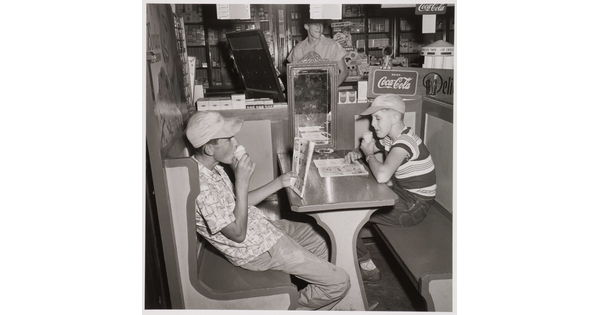 A black-and-white photograph of two White boys wearing baseball hats and eating ice cream cones in a booth.