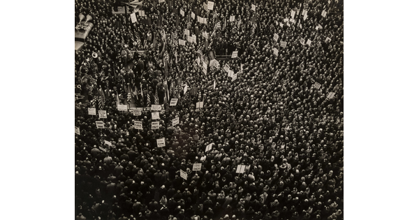 A black-and-white aerial photograph of a crowd of people, some carrying signs and American flags.