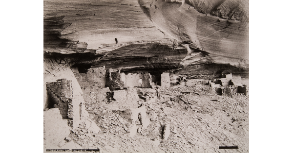 A black-and-white photograph of adobe ruins under an overhanging cliff ledge.
