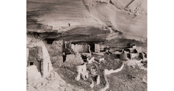 A black-and-white photograph of adobe ruins under an overhanging cliff ledge.