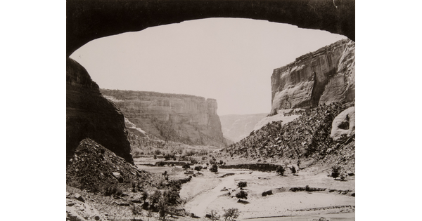 A black-and-white photograph of a canyon landscape framed by an arched rock overhang.