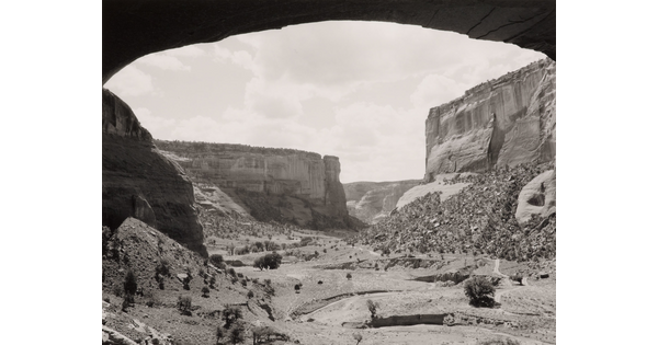 A black-and-white photograph of a canyon landscape framed by an arched rock overhang.