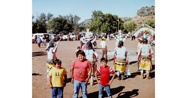 A color photograph of a three boys posing as a Native American dance takes place behind them.