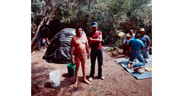 A color photograph of two medium-skinned men: one man wearing a loin cloth and the other in jeans and a t-shirt standing in a wooded setting.