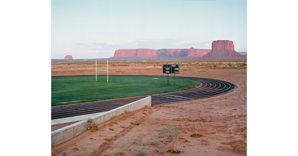 A color photograph of an athletic track and field with sunlit mesas and buttes in the distance.