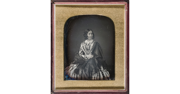 A black-and-white portrait photograph in a gold-frame of a White woman with her hair styled in ringlets, wearing a plaid dress with a full skirt and a shawl wrapped around her shoulders.