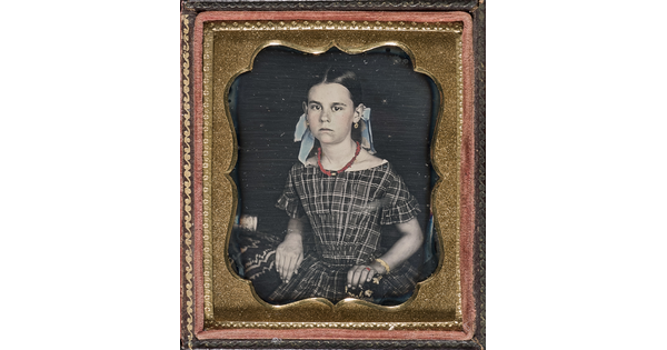 A hand-colored photograph in a decorative gold frame of a young White girl seated, wearing a plaid dress and bows in her hair.