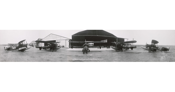 A black-and-white panoramic photograph of an airplane hanger with five biplanes in the foreground.