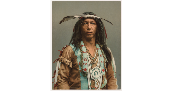 A colored photograph portrait of an Indigenous man in traditional clothing, including beaded necklaces and large feathers on his head.