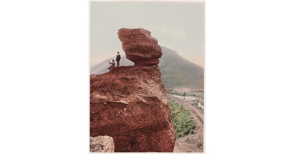 A color photograph of two men, one seated and one standing, next to a large rock formation with a mountain in the background.