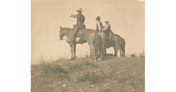 A sepia-toned photograph of three cowboys, one gesturing to the distance, on horseback in a field.