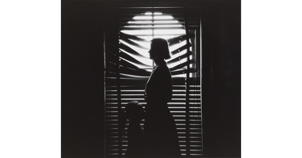 A black-and-white photograph of the silhouettes of a woman and young girl facing each other in front of a window with light coming through partially closed blinds.