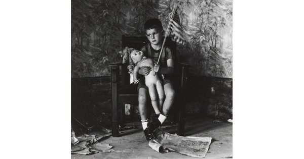 A black-and-white photograph of a young White boy in a chair holding a naked doll on his lap and waving an American flag.