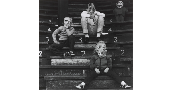 A black-and-white photograph of four children in grotesque costume masks sitting on wooden steps.