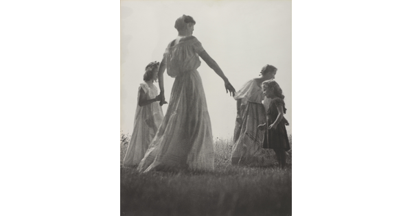 A black-and-white photograph of White women and children wearing long dresses holding hands to form a circle in a grassy field.