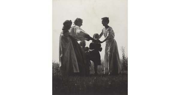 A black-and-white photograph of White women and children wearing long dresses holding hands to form a circle in a grassy field.
