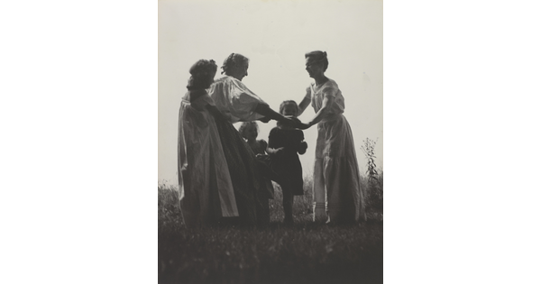 A black-and-white photograph of White women and children wearing long dresses holding hands to form a circle in a grassy field.