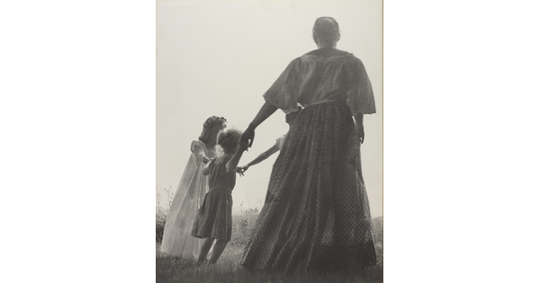A black-and-white photograph of a woman, seen from the back, and two White children holding hands in a grassy field.