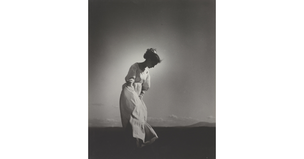 A black-and-white photograph of a White woman wearing a flowing white dress and looking down as she stands in a field.
