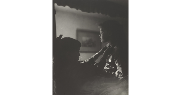 A black-and-white photograph of two young White girls, one fastening a ribbon around the neck of the other, both in shadow.