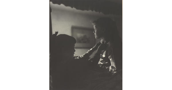 A black-and-white photograph of two young White girls, one fastening a ribbon around the neck of the other, both in shadow.