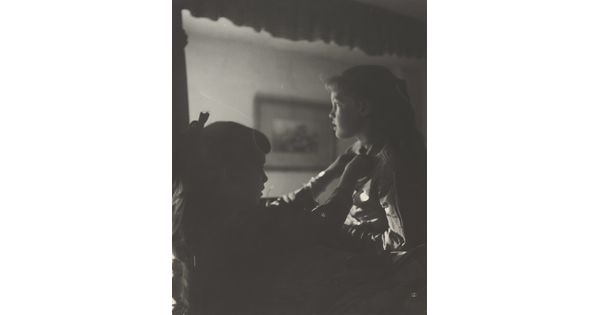 A black-and-white photograph of two young White girls, one fastening a ribbon around the neck of the other, both in shadow.