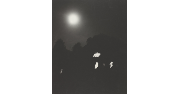 A black-and-white photograph of trees and a dark house with lit windows at night under a full moon.