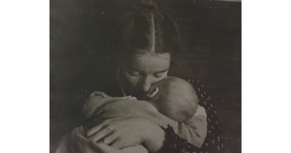 A black-and-white photograph of a White woman holding an infant to her chest while she rests her cheek on the infant's cheek.
