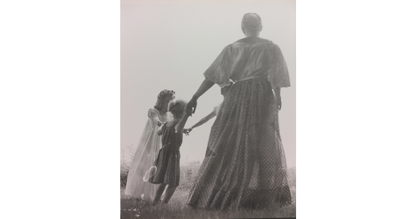 A black-and-white photograph of a woman, seen from the back, and two White children holding hands in a grassy field.