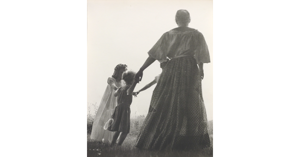 A black-and-white photograph of a woman, seen from the back, and two White children holding hands in a grassy field.