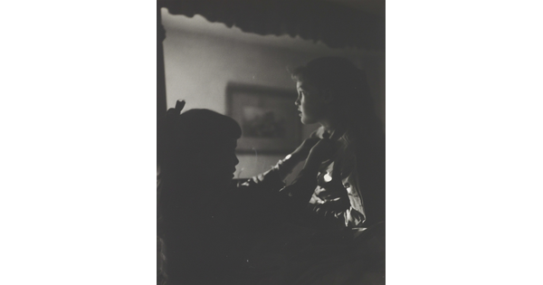 A black-and-white photograph of two young White girls, one fastening a ribbon around the neck of the other, both in shadow.