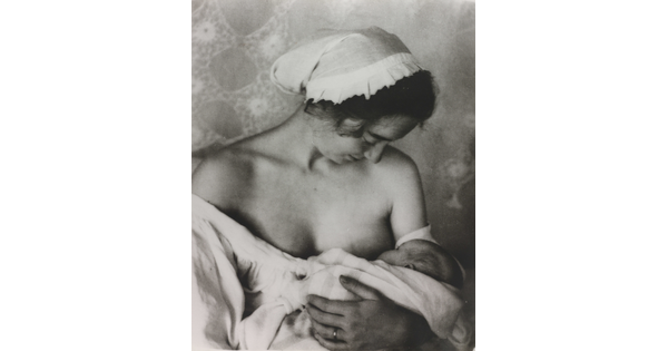 A black-and-white photograph of a White woman in a nightcap and white flowing blouse nursing an infant.