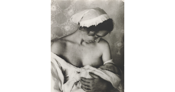 A black-and-white photograph of a White woman in a nightcap and white flowing blouse nursing an infant.