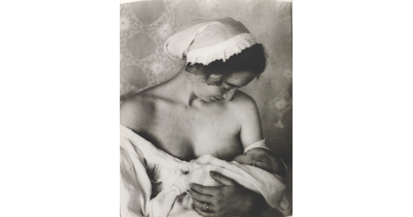 A black-and-white photograph of a White woman in a nightcap and white flowing blouse nursing an infant.