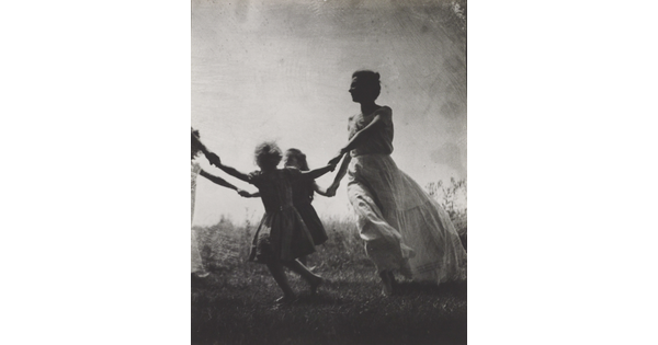 A black-and-white photograph of a White woman and children wearing long dresses holding hands to form a circle in a grassy field.