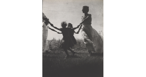 A black-and-white photograph of a White woman and children wearing long dresses holding hands to form a circle in a grassy field.